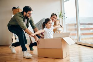 Happy little girl with arms outstretched having fun being pushed by her parents in carton box at new home